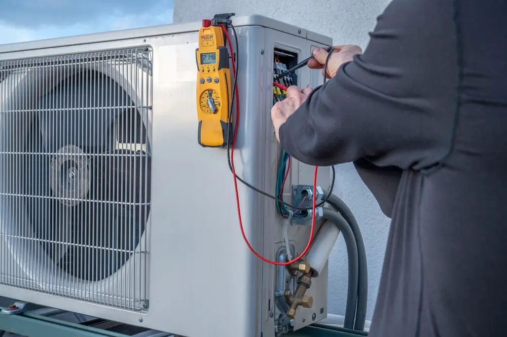 Multimeter on mini-split, hands testing voltage with test leads at power terminals.