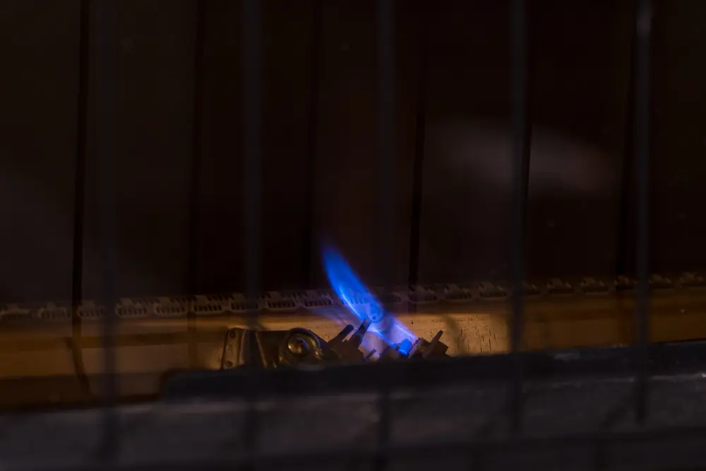 Small pilot flame for gas furnace heater. Constant blue flame for igniting main burners, behind safety grill.